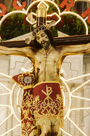 A religious statue depicts Jesus on the cross, adorned with an intricate golden crown and wearing a richly decorated red cloth with gold embroidery. The background features illuminated circular patterns and hints of greenery and flowers.