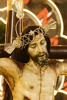 A realistic and detailed sculpture of Jesus on the cross, featuring a crown of thorns with radiant, decorative elements. The expression on the face is serene yet solemn, with closed eyes and a gentle tilt of the head. There are visible wounds and blood marks on the body, emphasizing the crucifixion. The background includes glowing neon lights that add a modern contrast to the traditional religious imagery.