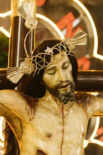 A realistic and detailed sculpture of Jesus on the cross, featuring a crown of thorns with radiant, decorative elements. The expression on the face is serene yet solemn, with closed eyes and a gentle tilt of the head. There are visible wounds and blood marks on the body, emphasizing the crucifixion. The background includes glowing neon lights that add a modern contrast to the traditional religious imagery.