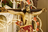 A depiction of Jesus Christ on the cross, adorned with a crown of thorns. The figure is wearing a red cloth with intricate gold embroidery around the waist. The background includes bright neon lights and green foliage, contributing to a dramatic and spiritual ambiance.
