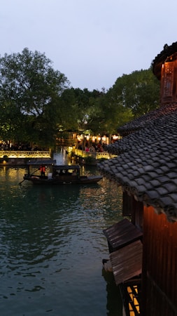 A traditional wooden boat is calmly gliding on a tranquil river lined with lush green trees. On the side, an ancient building with a tiled roof is visible, reflecting the warm lights from within onto the water surface. The scene is illuminated by soft lighting, giving the place a serene and picturesque atmosphere.