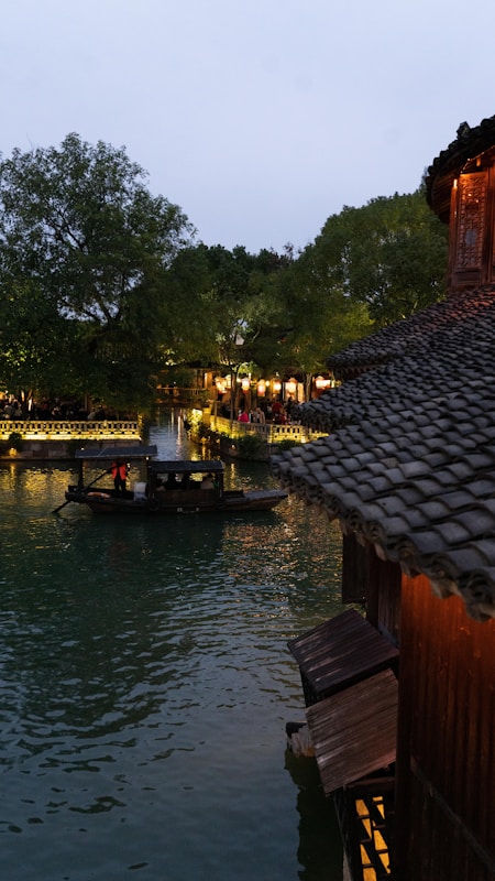 A traditional wooden boat is calmly gliding on a tranquil river lined with lush green trees. On the side, an ancient building with a tiled roof is visible, reflecting the warm lights from within onto the water surface. The scene is illuminated by soft lighting, giving the place a serene and picturesque atmosphere.