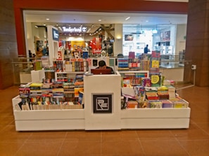 A bookstore kiosk in a shopping mall corridor is stacked with various books on all sides. A person is seated at the central counter with a laptop. Shops are visible in the background, including some with sale signs.