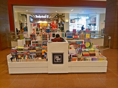 A bookstore kiosk in a shopping mall corridor is stacked with various books on all sides. A person is seated at the central counter with a laptop. Shops are visible in the background, including some with sale signs.