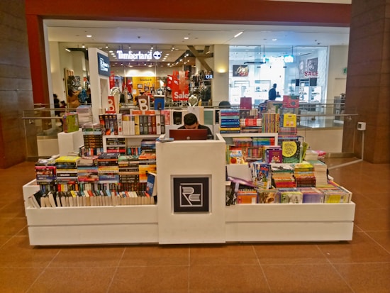 A bookstore kiosk in a shopping mall corridor is stacked with various books on all sides. A person is seated at the central counter with a laptop. Shops are visible in the background, including some with sale signs.