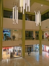 A spacious mall interior featuring multiple floors with a modern design. Large cylindrical lights hang from the high ceiling, illuminating the area. The ground floor consists of various retail stores with glass fronts displaying clothes and sale signs. A few people are walking and shopping throughout the mall.