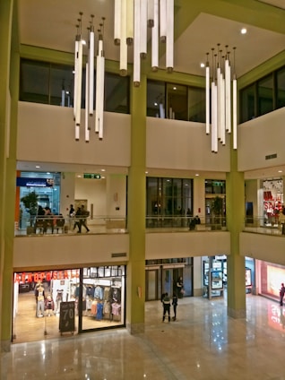 A spacious mall interior featuring multiple floors with a modern design. Large cylindrical lights hang from the high ceiling, illuminating the area. The ground floor consists of various retail stores with glass fronts displaying clothes and sale signs. A few people are walking and shopping throughout the mall.