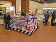 A candy and snacks kiosk in a shopping mall features a variety of colorful candies and packaged snacks. A customer, wearing a scarf and carrying a shopping bag, is selecting items from the display while the vendor, dressed in a green uniform, stands on the opposite side. Other shoppers are visible in the background, walking past the kiosk.