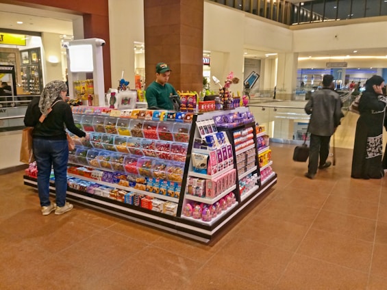 A candy and snacks kiosk in a shopping mall features a variety of colorful candies and packaged snacks. A customer, wearing a scarf and carrying a shopping bag, is selecting items from the display while the vendor, dressed in a green uniform, stands on the opposite side. Other shoppers are visible in the background, walking past the kiosk.