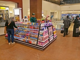 A candy and snacks kiosk in a shopping mall features a variety of colorful candies and packaged snacks. A customer, wearing a scarf and carrying a shopping bag, is selecting items from the display while the vendor, dressed in a green uniform, stands on the opposite side. Other shoppers are visible in the background, walking past the kiosk.