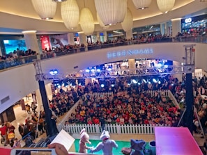 A crowded shopping mall with a large gathering of people watching a live performance. Two performers in animal costumes are on a small stage surrounded by a white picket fence. The audience, primarily children, is seated on the floor, while additional spectators stand around the fence and on the upper balcony levels. Bright stage lights illuminate the area, and several large decorative ceiling lamps hang above.