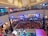 A crowded shopping mall with a large gathering of people watching a live performance. Two performers in animal costumes are on a small stage surrounded by a white picket fence. The audience, primarily children, is seated on the floor, while additional spectators stand around the fence and on the upper balcony levels. Bright stage lights illuminate the area, and several large decorative ceiling lamps hang above.