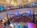 A crowded shopping mall with a large gathering of people watching a live performance. Two performers in animal costumes are on a small stage surrounded by a white picket fence. The audience, primarily children, is seated on the floor, while additional spectators stand around the fence and on the upper balcony levels. Bright stage lights illuminate the area, and several large decorative ceiling lamps hang above.