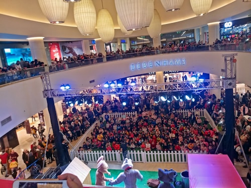 A crowded shopping mall with a large gathering of people watching a live performance. Two performers in animal costumes are on a small stage surrounded by a white picket fence. The audience, primarily children, is seated on the floor, while additional spectators stand around the fence and on the upper balcony levels. Bright stage lights illuminate the area, and several large decorative ceiling lamps hang above.