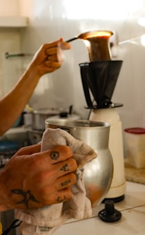 A person is preparing coffee using a traditional cloth filter. The process involves pouring hot water over coffee grounds contained in the cloth filter, which is held by a metal stand. The individual holds a spoon and a small jug, with visible tattoos on their hand.