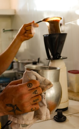 A person is preparing coffee using a traditional cloth filter. The process involves pouring hot water over coffee grounds contained in the cloth filter, which is held by a metal stand. The individual holds a spoon and a small jug, with visible tattoos on their hand.