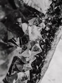 A close-up view of a cluster of jagged, geometric crystal formations with a mixture of translucent and reflective surfaces. The crystals create an intricate pattern and have a monochromatic grayscale tone.