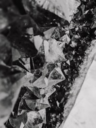 A close-up view of a cluster of jagged, geometric crystal formations with a mixture of translucent and reflective surfaces. The crystals create an intricate pattern and have a monochromatic grayscale tone.