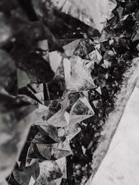 A close-up view of a cluster of jagged, geometric crystal formations with a mixture of translucent and reflective surfaces. The crystals create an intricate pattern and have a monochromatic grayscale tone.