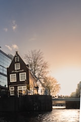 A picturesque canal scene featuring a traditional gabled house with the name 'De Stuyveracht' on its facade. The building is situated next to a waterway, bathed in the warm hues of a setting sun. Leafless trees and a modern glass building are also visible.