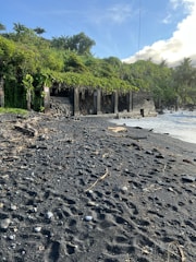 A black sand beach with various driftwood and small stones scattered across the sand. There are several concrete structures with vines and other vegetation growing over them, surrounded by dense greenery including trees and bushes. The ocean waves gently reach the shore, and the sky is mostly clear with a few clouds.