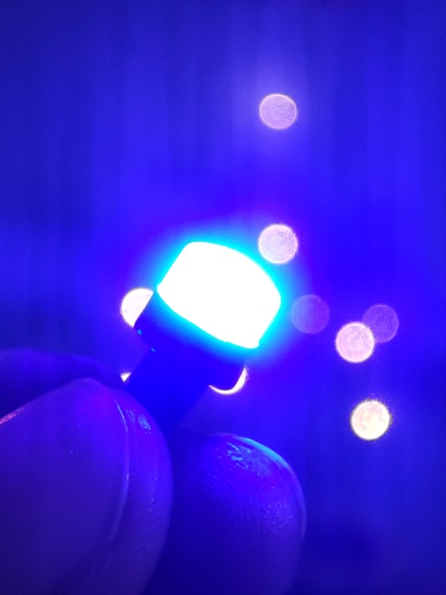 A close-up of a hand holding a small, glowing LED light that emits a bright blue color. In the background, bokeh lights create soft circles of varying intensity, enhancing the image's depth.