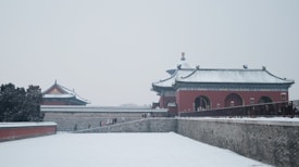 Traditional Chinese architecture stands covered in snow, with muted colors and intricate roof designs. A few people walk along the path in the wintry scene, surrounded by historic buildings and neatly manicured trees.