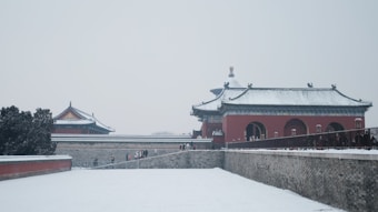 Traditional Chinese architecture stands covered in snow, with muted colors and intricate roof designs. A few people walk along the path in the wintry scene, surrounded by historic buildings and neatly manicured trees.
