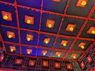 A decorative ceiling featuring a grid-like pattern with recessed squares. Each square contains a light fixture surrounded by red frilled fabric and plaid patterns. The ceiling is predominantly black with highlights of red, yellow, and blue throughout.