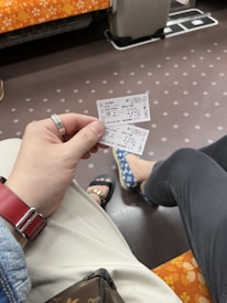 A hand holding two train tickets inside a train compartment. The floor is patterned with small dots, and the seat covers have an orange and white floral design. A red watch strap is visible on the wrist, and the person is wearing light-colored pants. Another person is seated nearby, wearing dark pants and blue-patterned shoes.