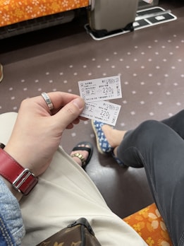 A hand holding two train tickets inside a train compartment. The floor is patterned with small dots, and the seat covers have an orange and white floral design. A red watch strap is visible on the wrist, and the person is wearing light-colored pants. Another person is seated nearby, wearing dark pants and blue-patterned shoes.