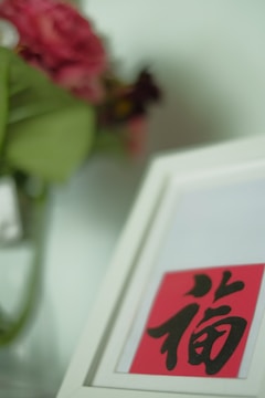 A close-up of a white-framed image featuring Chinese calligraphy on a red background. In the blurred background, there is a vase with pink and red flowers.