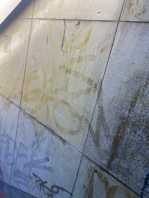 The image depicts a wall of tiles covered with faded graffiti in yellow and black. The tiles show signs of wear and age, with visible cracks and weathering. The graffiti is abstract and features a mix of lines, shapes, and letters.