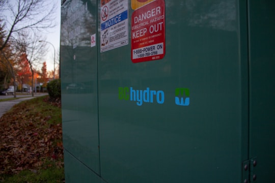 A large green electrical utility box is situated outdoors, with several warning signs attached to it, including one for electrical hazards. The BC Hydro logo is prominently displayed. Fallen autumn leaves are scattered on the grass nearby, and there are trees with autumn foliage along a sidewalk in the background.
