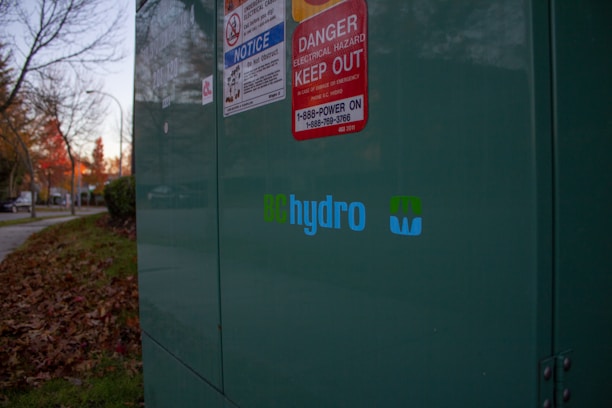 A large green electrical utility box is situated outdoors, with several warning signs attached to it, including one for electrical hazards. The BC Hydro logo is prominently displayed. Fallen autumn leaves are scattered on the grass nearby, and there are trees with autumn foliage along a sidewalk in the background.
