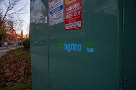 A large green electrical utility box is situated outdoors, with several warning signs attached to it, including one for electrical hazards. The BC Hydro logo is prominently displayed. Fallen autumn leaves are scattered on the grass nearby, and there are trees with autumn foliage along a sidewalk in the background.