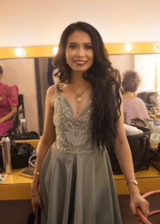 A woman with long, dark hair is dressed in an elegant, grey gown with detailed embroidery. She is standing in a well-lit dressing room, smiling directly at the camera. The background includes a brightly lit mirror, some cosmetics, and another person seated, partially visible.