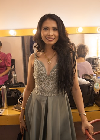 A woman with long, dark hair is dressed in an elegant, grey gown with detailed embroidery. She is standing in a well-lit dressing room, smiling directly at the camera. The background includes a brightly lit mirror, some cosmetics, and another person seated, partially visible.