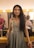 A woman with long, dark hair is dressed in an elegant, grey gown with detailed embroidery. She is standing in a well-lit dressing room, smiling directly at the camera. The background includes a brightly lit mirror, some cosmetics, and another person seated, partially visible.