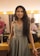 A woman with long, dark hair is dressed in an elegant, grey gown with detailed embroidery. She is standing in a well-lit dressing room, smiling directly at the camera. The background includes a brightly lit mirror, some cosmetics, and another person seated, partially visible.