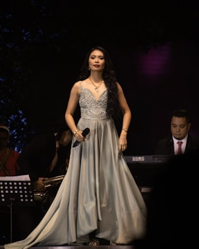 A woman in a flowing, elegant gown holds a microphone on stage. She is wearing jewelry and her long hair cascades over her shoulders. There are musicians in the background, with one playing a keyboard and another holding a saxophone.