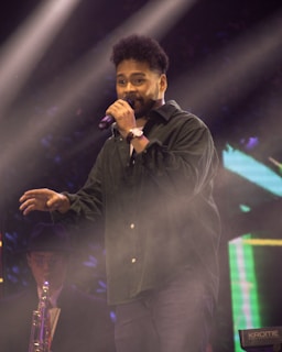 A person is performing on stage with a microphone in hand. They are wearing a dark shirt and jeans. In the background, another musician is playing a saxophone and wearing a hat and glasses. The stage is illuminated with bright lights, creating a lively and vivid atmosphere.
