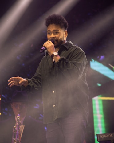 A person is performing on stage with a microphone in hand. They are wearing a dark shirt and jeans. In the background, another musician is playing a saxophone and wearing a hat and glasses. The stage is illuminated with bright lights, creating a lively and vivid atmosphere.