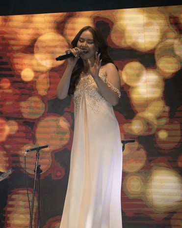A woman in a white gown is singing passionately on stage, holding a microphone. She stands in front of a vivid, abstract background with swirling patterns and warm colors. The stage setting includes another microphone on a stand visible beside her.