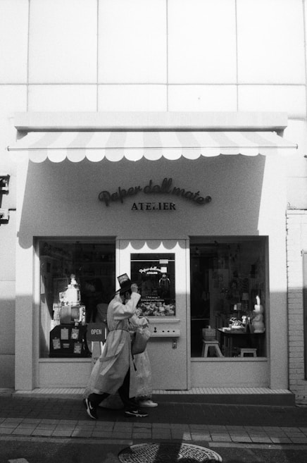 A person dressed in a long coat and hat is walking past a shop with a striped awning. The shop, named 'Atelier', appears to be connected to the brand 'Paper doll mate'. Inside the window display, various items are present, including an 'OPEN' sign. The scene is captured in black and white, adding a nostalgic or vintage feel.
