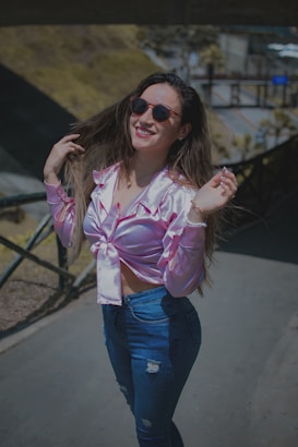 A woman smiling while posing outdoors in a casual outfit. She is wearing a pink satin blouse, blue jeans, and sunglasses. Her long hair flows freely as she holds it with one hand, and the background features a blurred outdoor setting with pathways and greenery.