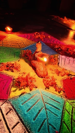 A vibrant and intricate rangoli design on the floor, featuring a colorful geometric pattern with shades of blue, green, yellow, pink, and orange. In the center, there is a small clay figurine with a lit candle inside, surrounded by marigold petals. Additional oil lamps add to the warm, glowing ambiance.