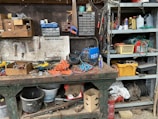 A cluttered workshop with various tools and equipment scattered across a workbench. Industrial items such as gloves, a welder, a can of lubricant spray, and multiple boxes with drawers filled with screws and hardware are visible. Shelves are filled with assorted items including ropes, containers, and miscellaneous repair supplies. The scene conveys a busy and industrious environment.