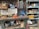 A cluttered workshop with various tools and equipment scattered across a workbench. Industrial items such as gloves, a welder, a can of lubricant spray, and multiple boxes with drawers filled with screws and hardware are visible. Shelves are filled with assorted items including ropes, containers, and miscellaneous repair supplies. The scene conveys a busy and industrious environment.