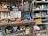 A cluttered workshop with various tools and equipment scattered across a workbench. Industrial items such as gloves, a welder, a can of lubricant spray, and multiple boxes with drawers filled with screws and hardware are visible. Shelves are filled with assorted items including ropes, containers, and miscellaneous repair supplies. The scene conveys a busy and industrious environment.